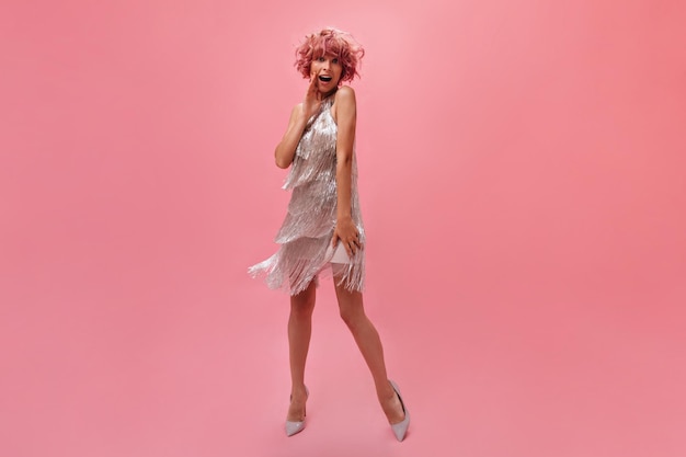 Surprised happy woman in silver shiny dress and high heels poses on isolated Curly young girl with tattoos on arm looks shocked on pink background