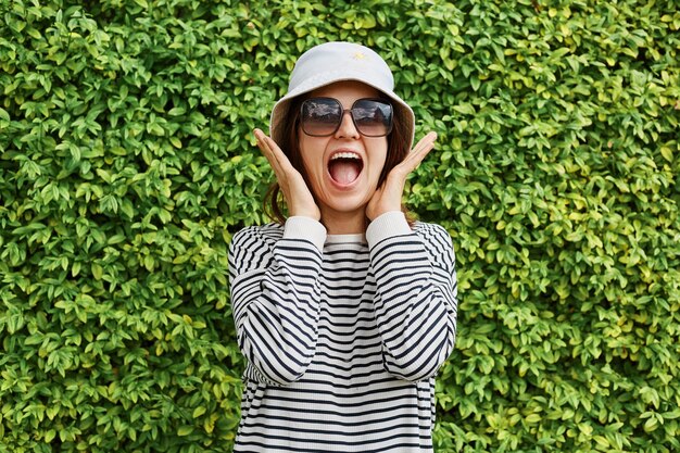 Surprised happy woman wearing striped shirt panama and sunglasses standing against green garden fence screaming with happiness enjoying summertime