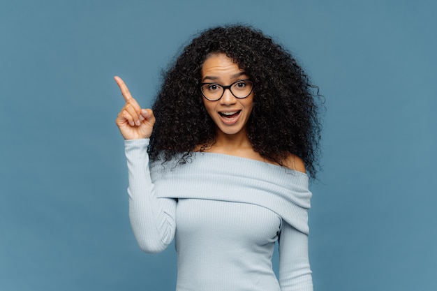 Surprised happy woman with Afro hairstyle, points fore finger on blank space above, has opened mouth