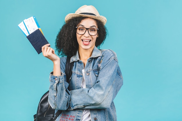スタジオで青色の背景に分離された驚いて幸せな若いアフリカ系アメリカ人の女の子。旅行の概念。パスポート、搭乗券のチケットを保持します。