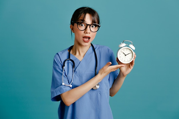 Surprised holding alarm clock young female doctor wearing uniform fith stethoscope isolated on blue background