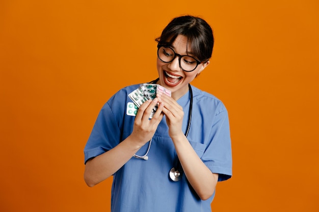 Surprised holding pills young female doctor wearing uniform fith stethoscope isolated on orange background