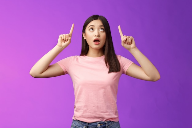 Surprised impressed asian girl brunette look fascinated up, drop jaw gasping astonished, check out impressive interesting promo, pointing top advertisement amazed, stand purple background.