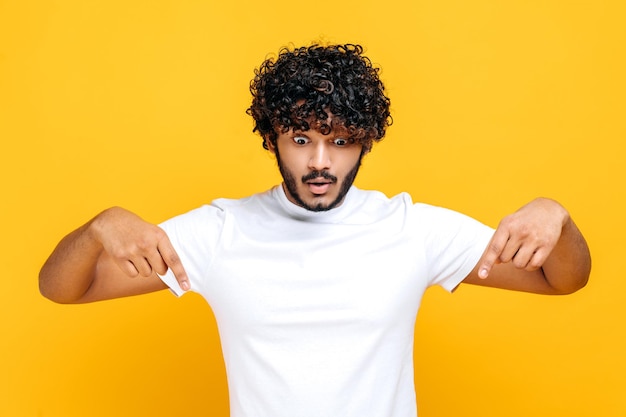 Photo surprised indian or arabian shocked curlyhaired guy in basic white tshirt amazed looks down and points fingers down excited face expression stands on isolated orange color background