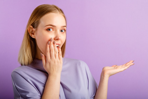 Surprised joyful excited blonde caucasian pretty young woman cover mouth with hand. Teenage girl show side gesture by hand on copy space place over isolated color purple background.