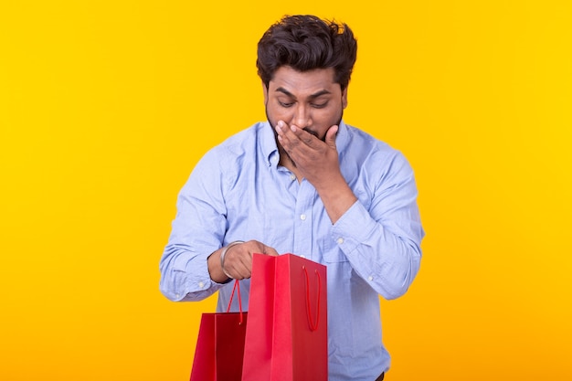 Surprised joyful young Indian man looks into the packages for shopping, standing on a yellow wall