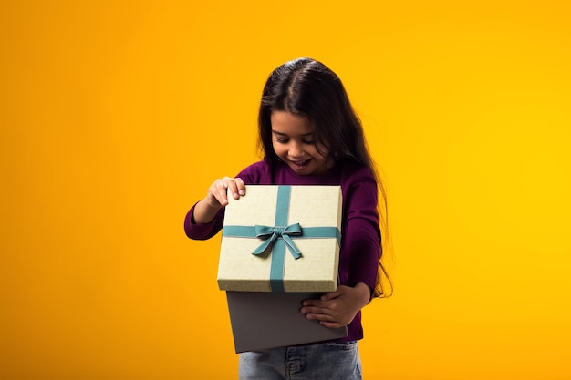 Foto ragazza sorpresa che tiene in mano una scatola regalo e guarda dentro la scatola su uno sfondo giallo concetto di compleanno e celebrazione