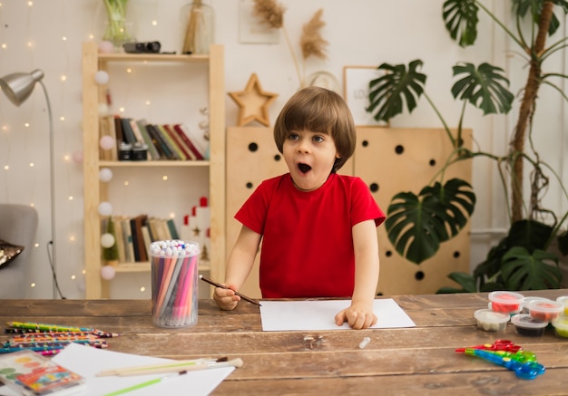 Surprised little boy draws with a colored felt-tip pen on white paper at a wooden desk with stationery in the room