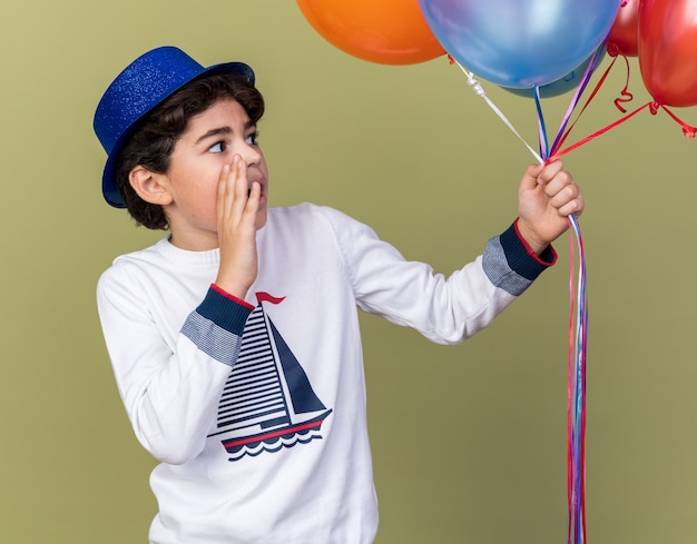 Surprised little boy wearing blue party hat holding and looking at balloons calling someone isolated on olive green wall