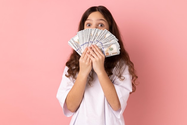 Surprised little girl covering half of face with big fan of dollar banknotes looking with big eyes