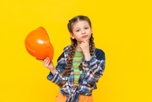 A surprised little girl holds a construction helmet The child chooses repairs in the nursery Yellow isolated background