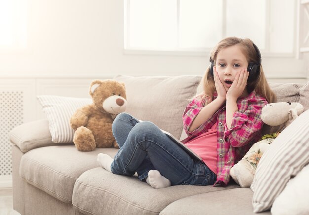 Surprised little girl playing online games on tablet and listening to music in headphones. Female child sitting on sofa with her teddy bear. Shocking content and social networking concept