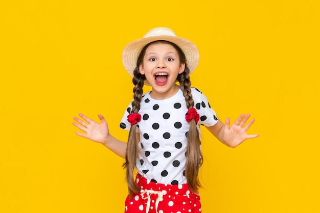 A surprised little girl in a straw hat and summer clothes with polka dots An astonished child on a yellow isolated background