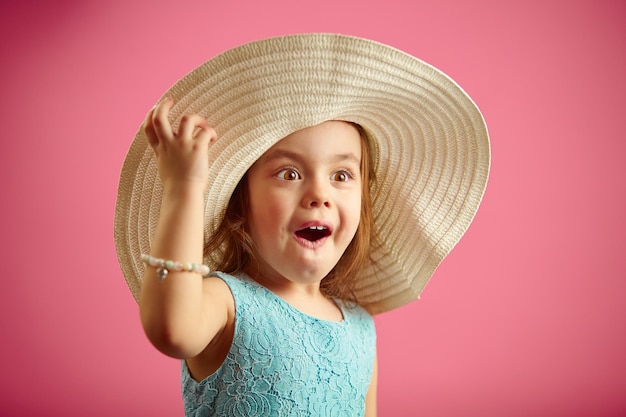 Surprised little girl wearing in straw beach hat having an amazement face stands on a pink isolated