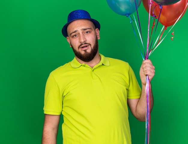 Surprised looking camera young man wearing blue party hat holding balloons 