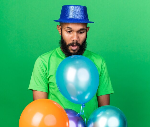 Surprised looking down young afro-american guy wearing party hat standing behind balloons 