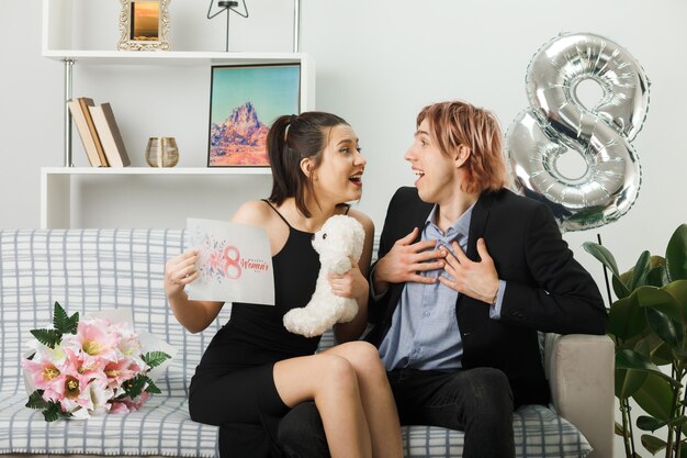 Surprised looking at each other young couple on happy women day with teddy bear and greeting card guy putting hands on heart sitting on sofa in living room