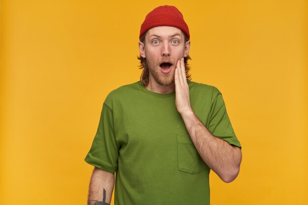 Surprised looking male, handsome bearded guy with blond hair. wearing green t-shirt and red beanie. has tattoos. touching face in amazement. isolated over yellow wall