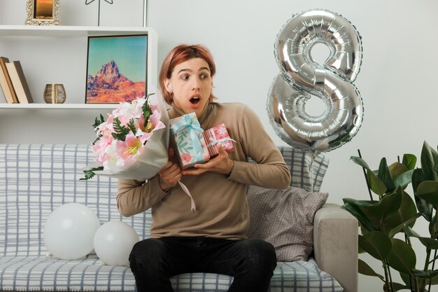 Surprised looking side handsome guy on happy women day holding present with bouquet sitting on sofa in living room