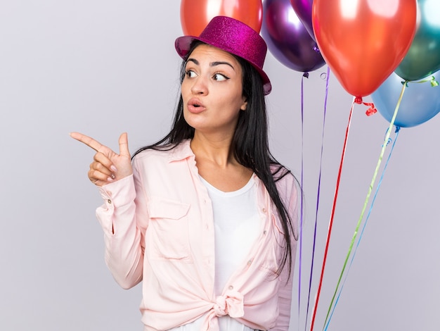 Surprised looking side young beautiful girl wearing party hat holding balloons and points at side isolated on white wall with copy space