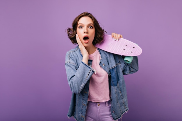 Surprised lovable woman in oversize jacket holding skateboard Indoor studio photo of debonair girl