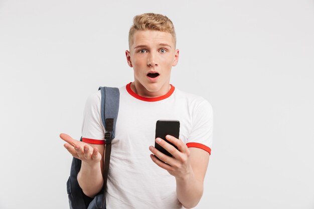 surprised male student having clean healthy skin wearing backpack using mobile phone and expressing puzzlement, isolated over white