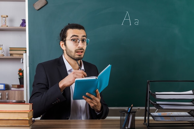 안경을 끼고 놀란 남자 교사는 교실에서 학교 도구를 가지고 탁자에 앉아 책에 글을 씁니다.