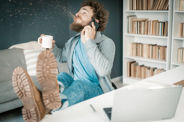 Photo surprised man hears the news on his smartphone and spits out coffee man has legs on the table and a ...