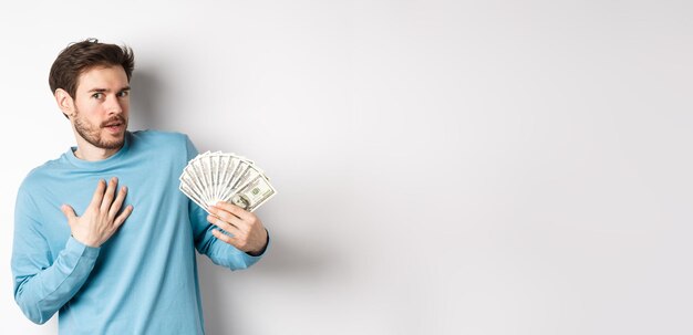 Photo surprised man holding money looking amazed at camera touching chest and gasping standing in blue swe