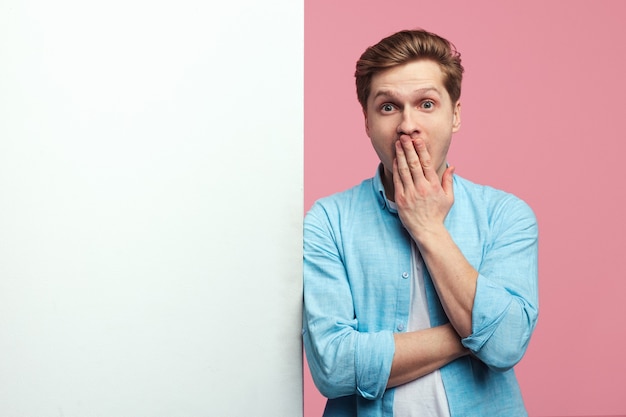 Surprised man standing alongside empty white billboard wall against pink