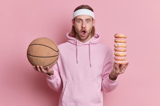 Surprised man in sweatshirt holds basketball and pile of sweet high calories doughnuts chooses between sport and unhealthy food keeps mmouth opened