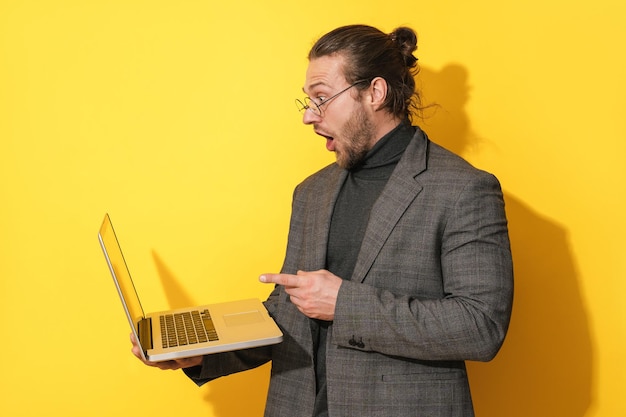 Surprised man wearing eyeglasses pointing at laptop screen on yellow background