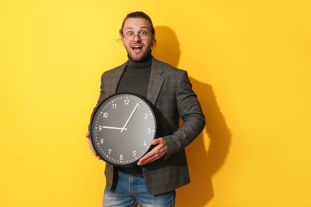 Surprised man wearing glasses holding big clock on yellow background