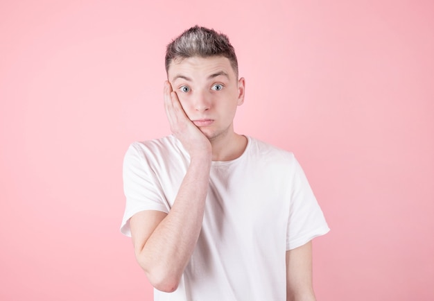 Surprised man in white t-shirt holding his face with hand on pink wall