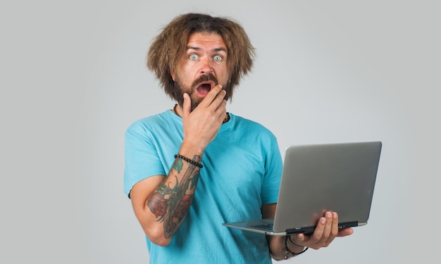 Удивленный мужчина с ноутбуком работает из дома, бородатый парень работает на компьютере фрилансером