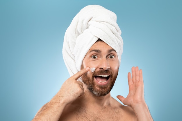 Surprised man with towel on head using face cream and looking at camera with excitement posing over