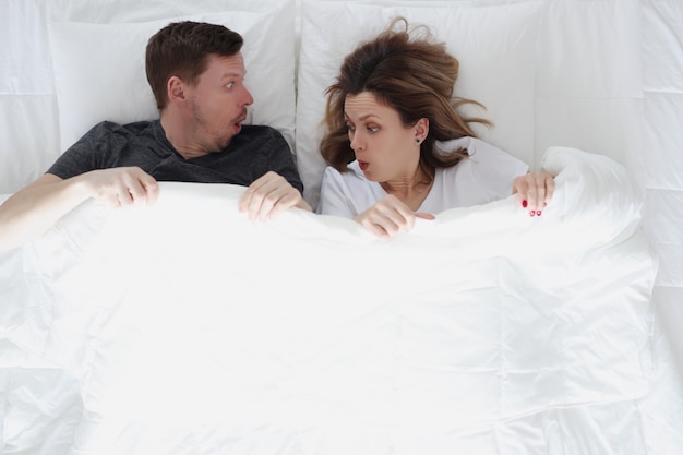 Surprised man and woman looking under covers while lying in bed. Morning erection in men concept