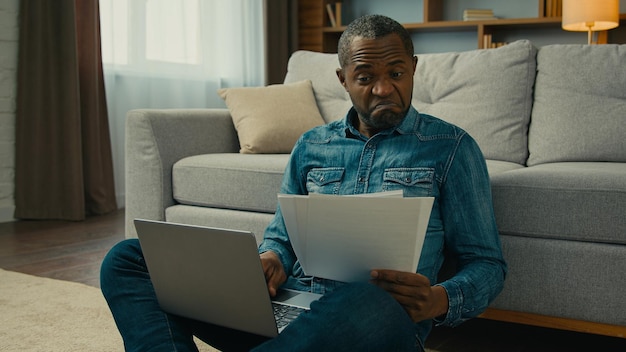 Surprised mature african american man enter data into laptop from paper documents happy with annual