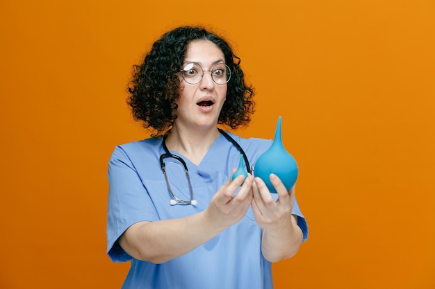 Surprised middleaged female doctor wearing uniform glasses and stethoscope around her neck stretching enemas out towards camera looking at them isolated on orange background