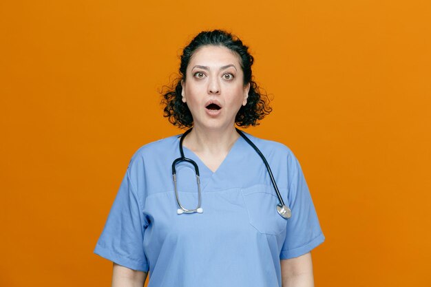 Medico femminile di mezza età sorpreso che indossa uniforme e stetoscopio intorno al collo guardando la fotocamera isolata su sfondo arancione