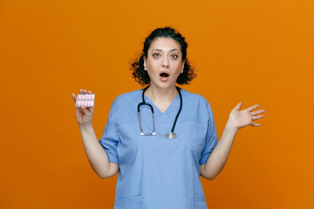 Surprised middleaged female doctor wearing uniform and stethoscope around her neck looking at camera showing pack of capsules and empty hand isolated on orange background