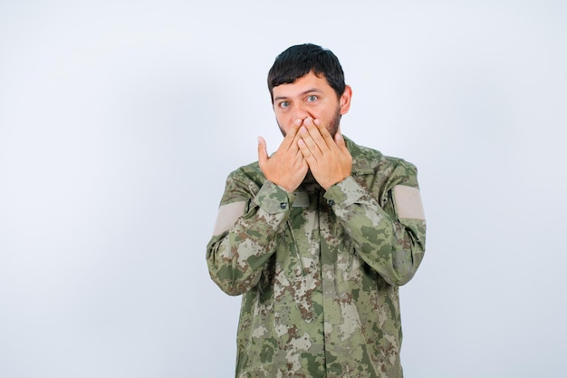 L'uomo militare sorpreso sta guardando la fotocamera tenendo le mani sulla bocca su sfondo bianco Foto Premium