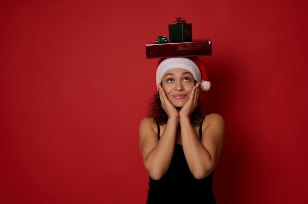 Surprised mysterious woman in Santa hat and evening black dress rejoicing looking up at Christmas gifts on her head, taking her hands on her cheeks, expressing happiness, isolated on red background