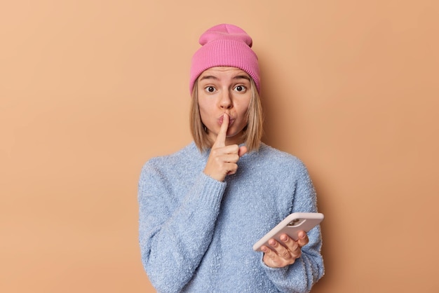 Surprised mysterious young woman makes shush gesture tells
secret holds mobile phone dressed in casual clothes asks to be
quiet isolated over brown background. hush dont say any word
please.