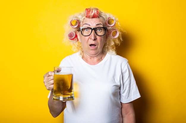 Surprised old woman with hair curlers with a glass of beer on a yellow surface