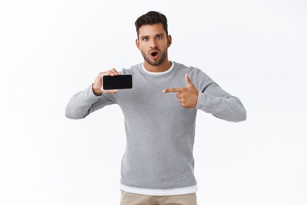 Surprised overwhelmed young handsome man in grey sweater asking question about mobile game or application, holding smartphone horizontally talking at camera intrigued, pointing screen