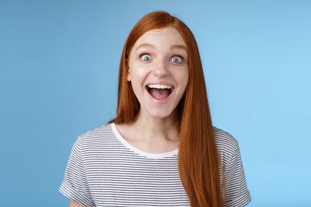 Surprised pleased happy impressed redhead european girl 20s reacting amused wide eyes look admiration joy receive incredible offer standing excited express thrill upbeat feelings, blue background.