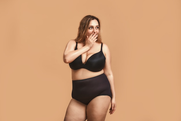 Surprised plump woman in black underwear covering her mouth with hand 