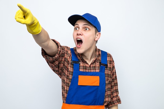 Surprised points at side young cleaning guy wearing uniform and cap with gloves 