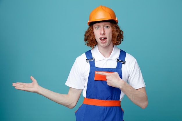 Surprised pretending holding and points at something young builder man in uniform isolated on blue background